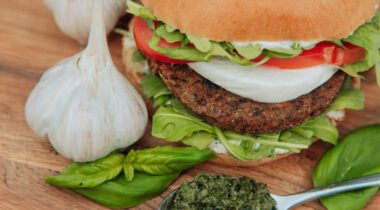 prepared garlic pesto burger on white bun with lettuce, tomato and onion. item on wood surface with whole garlic bulbs and spoonful of pesto sauce