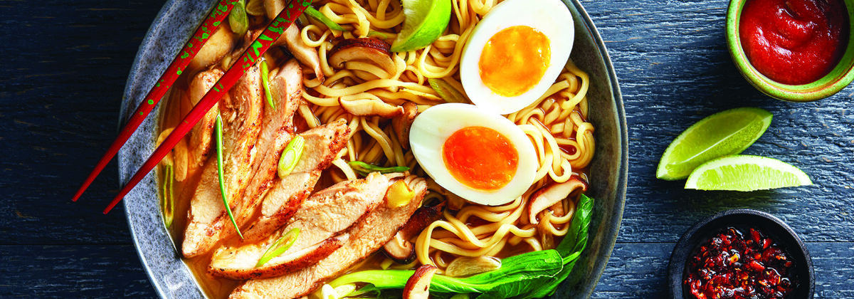 bowl of asian noodle soup with sliced chicken, cut hard boiled egg, shiitake mushroom stems. To the side are two lime wedges and two bowls of hot sauce.