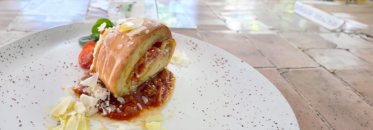 a speckled plate with a Pancetta Bread Roll in the middle on a bed of red sauce, sprinkles with Parmesan cheese.